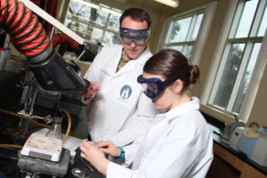 A man wearing a white lab coat and protective goggles looks at a woman wearing a white lab coat and protective goggles as she performs a scientific procedure in a lab. 