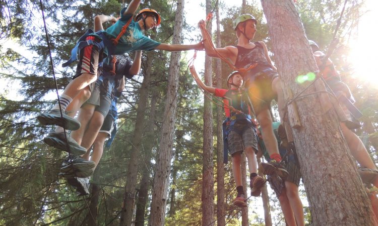 A group of teenagers wearing helmets on a ropes course in the woods. 
