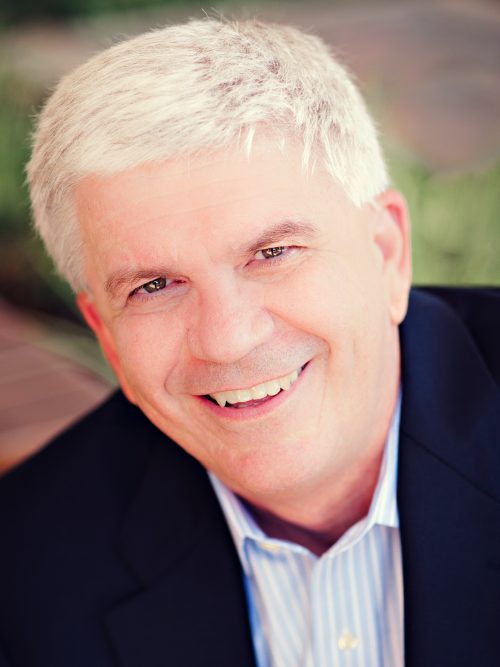 A man with short white hair wearing a blue striped shirt and blue suit jacket smiles for the camera. 