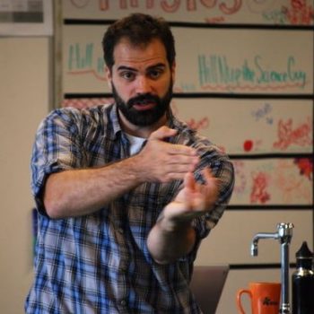 A man with dark hair and facial hair wearing a blue plaid shirt demonstrates something with his hands inside a classroom.