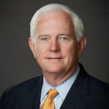 A man with white hair wearing a black suit and yellow tie smiles for the camera. 