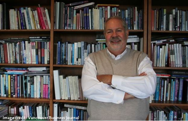 A man with gray hair and facial hear wearing a white shirt and tan sweater vest smiles for the camera in front of bookshelves. 