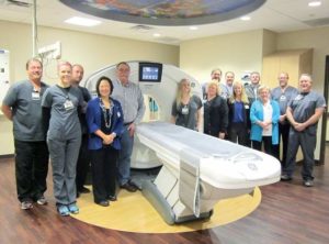 A group of people smile for the camera while gathered around CT-scanner. 