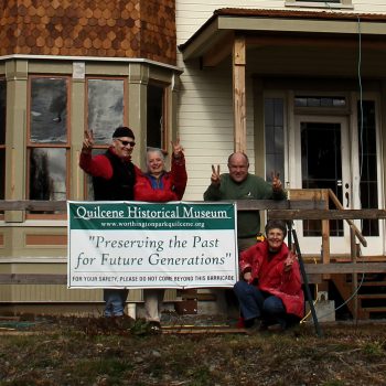 Four people smile in front of a historic building behind a sign that says "Quilcene Historical Museum: Preserving the Past for Future Generations"