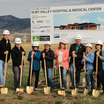 Eight people wearing hard hats and holding shovels smile for the camera in front of a sign that says "Future Home of the Ruby Valley Hospital & Medical Center." 