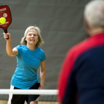 A woman with straight gray hair wearing a blue shirt plays pickleball on a pickleball court. 