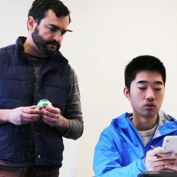 A man with short dark hair and facial hair wearing a dark vest and striped shirt looks at another man wearing a blue jacket looking at this phone. 