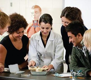 A group of adults gather around a woman wearing a white lab coat as she conducts an experiment. 