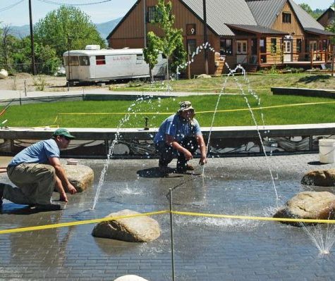 Two men wearing hats install a splash pad with sprinklers outdoors.