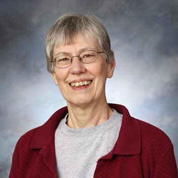 A woman with short gray hair wearing a gray shirt, a red jacket, and glasses smiles for the camera. 