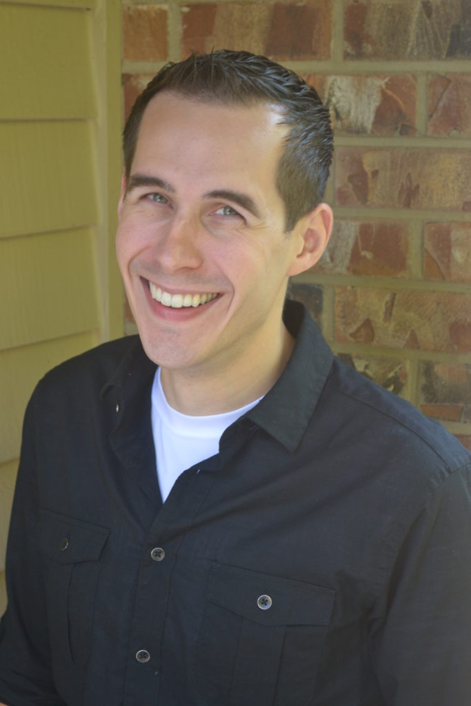 A man with short dark hair wearing a white shirt and black button-down smiles for the camera in front of a brick wall. 