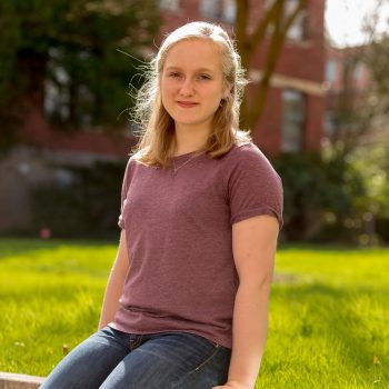 A young woman with straight blond hair wearing a purple tshirt and jeans sitting on a wall on a college campus. 