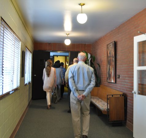 A group of adults walk through a hallway with brick walls on one side and windows on the other.