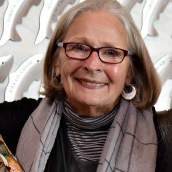 A woman with short gray hair wearing a gray scarf, silver earrings, and glasses smiles for the camera. 