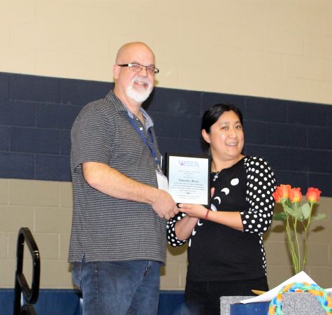 A man with white facial hair wearing glasses and a gray shirt holds a certificate with a woman with dark straight hair wearing a black dress.