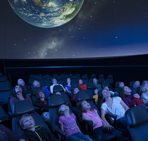 Students and adults look up at a projection of the earth inside a planetarium auditorium.
