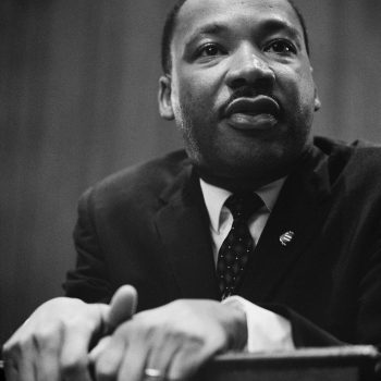 A photo of Martin Luther King, Jr. wearing a black suit, his hands on a podium in front of him. 