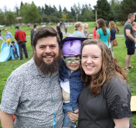 A man with dark hair and a beard and a woman with brown curly hair hold a child wearing a purple graduation cap and glasses and smile for the camera. They are outside on a lawn with many other people in the background behind them.