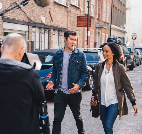 A man with short dark hair wearing a jeans jacket and a woman with dark curly hair wearing a brown jacket walk down a busy street talking to each other.