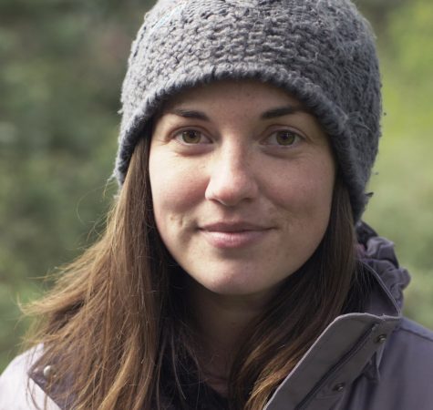 A woman with straight brown hair wearing a gray knit beanie looks at the camera.