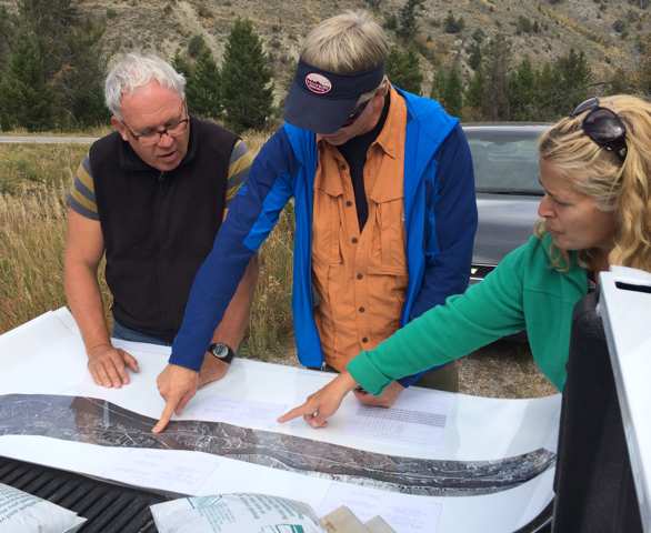 Three people analyze a large piece of paper and point to a map on it, while standing outside. 