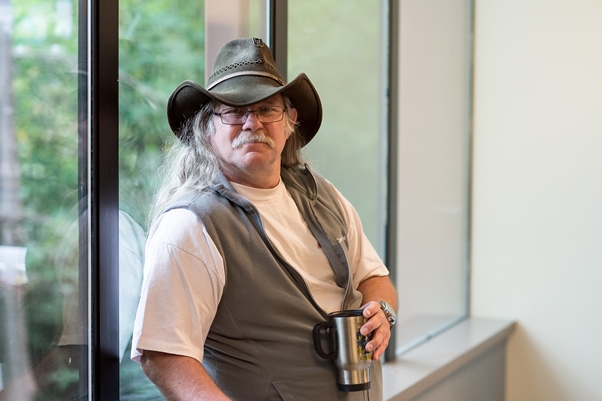 A man with long, straight gray hair and a mustache wearing a green cowboy hat, white t-shirt, and gray vest holding a coffee thermos looks at the camera near a window. 