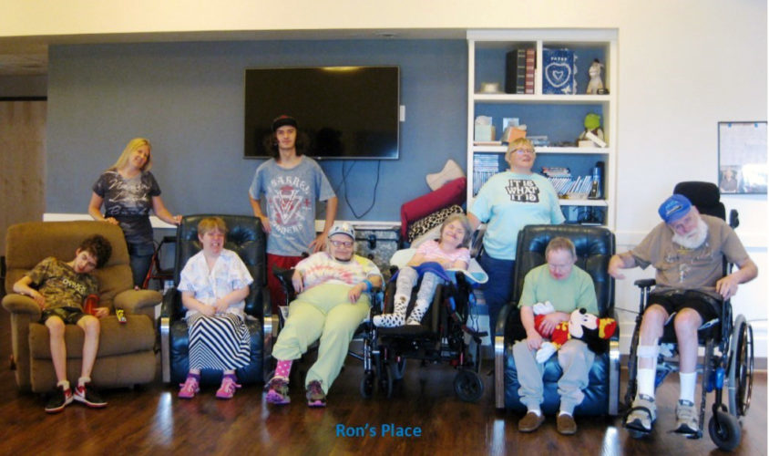 A group of people in wheelchairs or sitting in armchairs look at the camera with a TV and bookshelves behind them. 