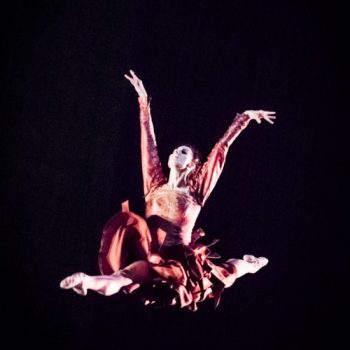 A ballerina wearing a pink dress performs a leap mid-air against a black background. 