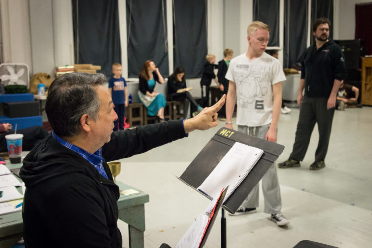 A man wearing a blue shirt and black jacket instructs actors in a studio. 