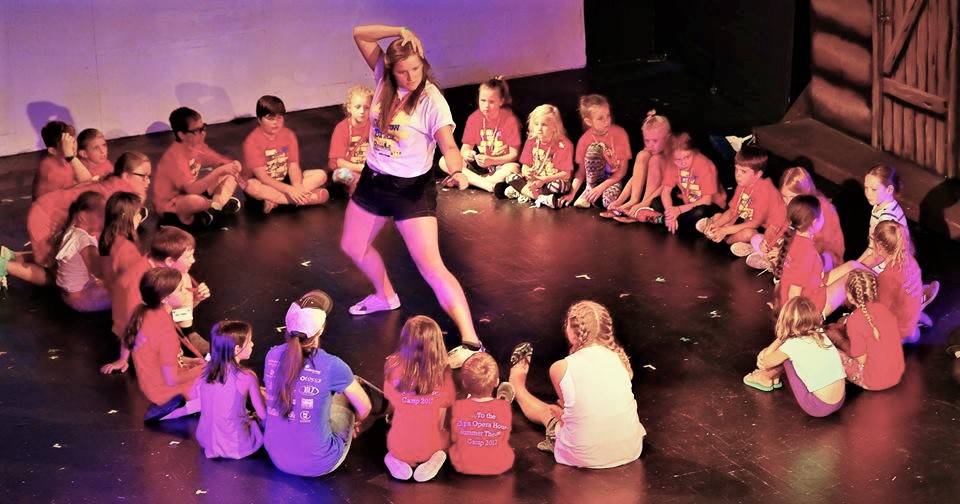 A young woman with long straight brown hair wearing a white t-shirt and black shorts acts out a scene in the middle of a group of young children on stage. 