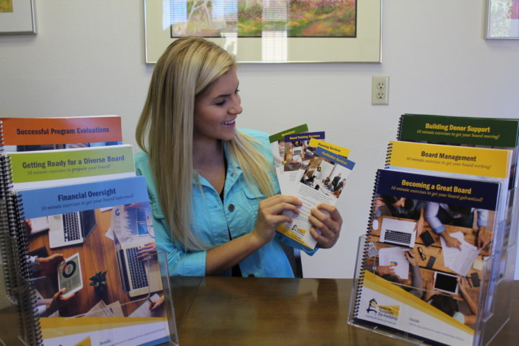 A woman with long, straight, blond hair smiles while holding pamphlets and sitting at a table. 