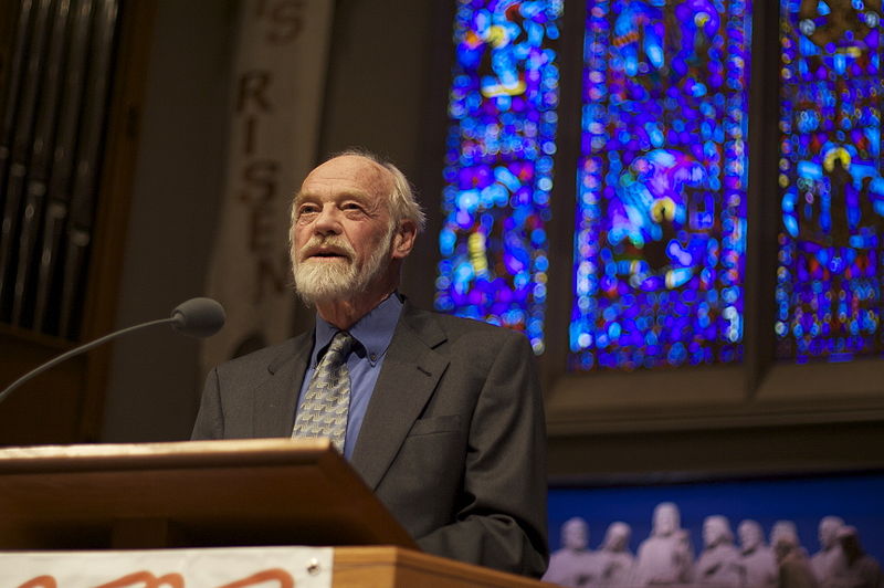 A man with white hair and a white beard wearing a dark gray suit speaks into a microphone at a podium with a stained glass window behind him. 