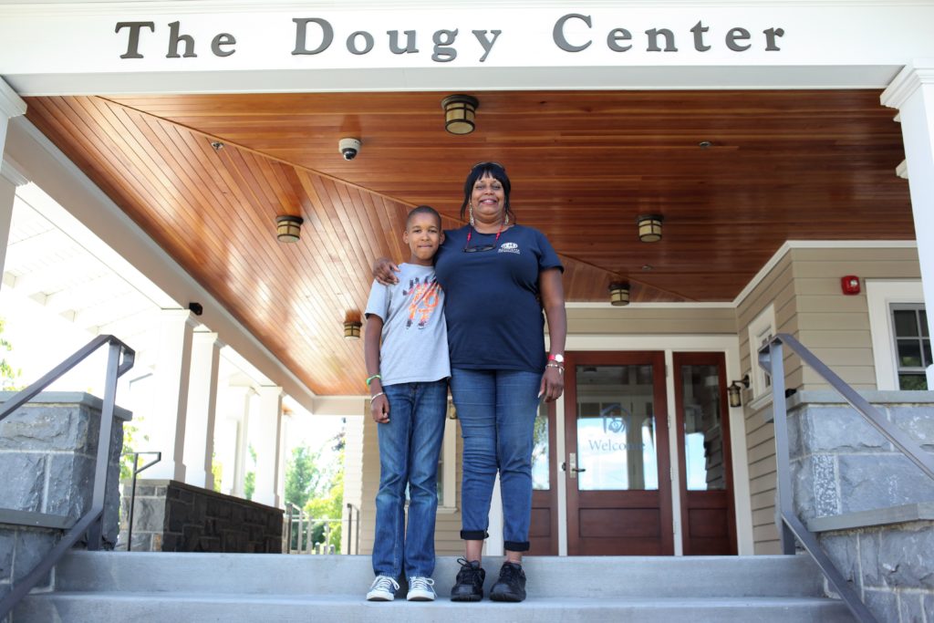 A woman wearing jeans and a blue t-shirt puts her arm around a boy wearing jeans and a gray t-shirt on the steps of a facility that says "The Dougy Center" above them. 