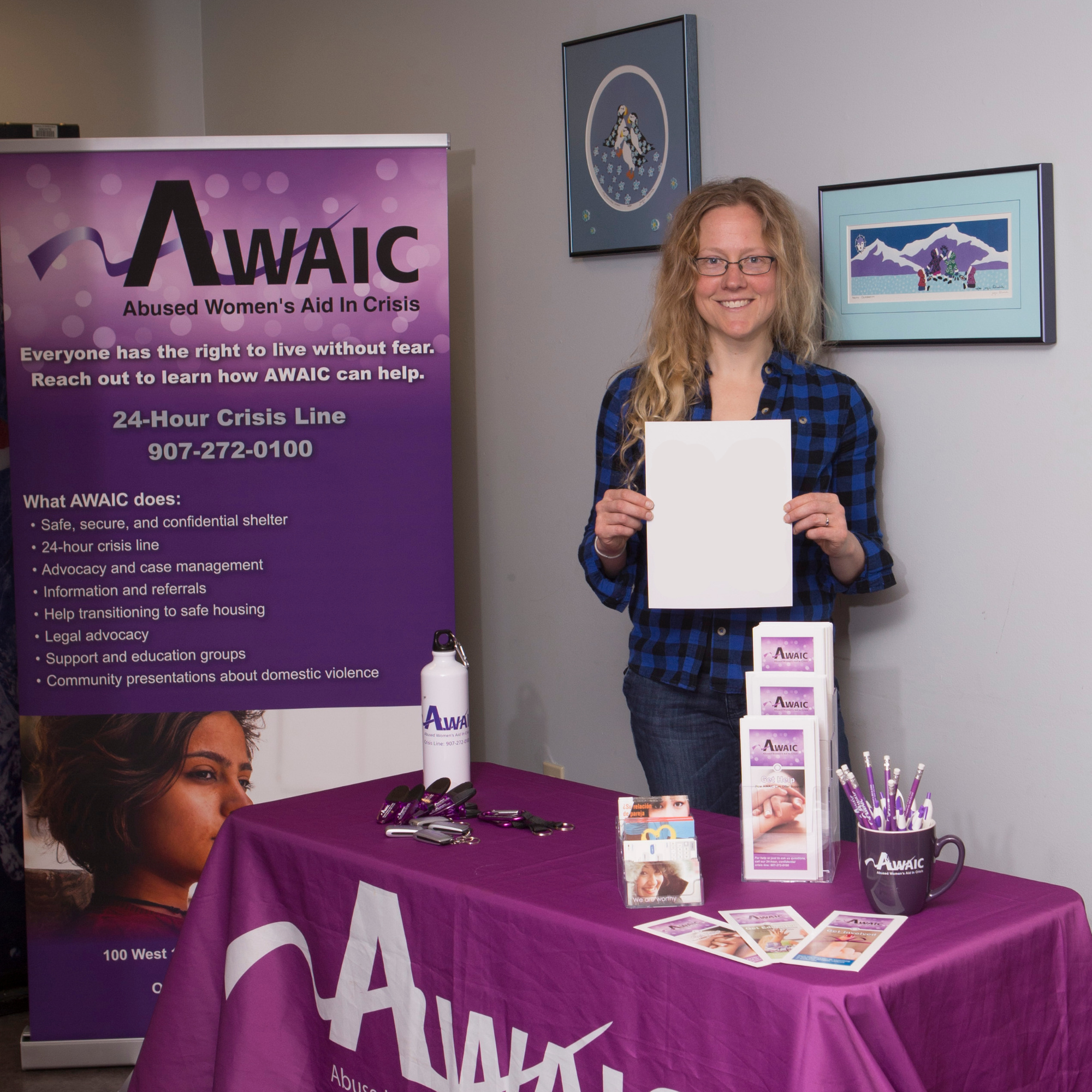 A woman with long blond hair wearing glasses and a blue plaid shirt smiles at the camera while holding a piece of paper. She stands behind a purple table with pens and brochures, next to a purple display with text that says "AWAIC: Abused Women's Aid in Crisis"