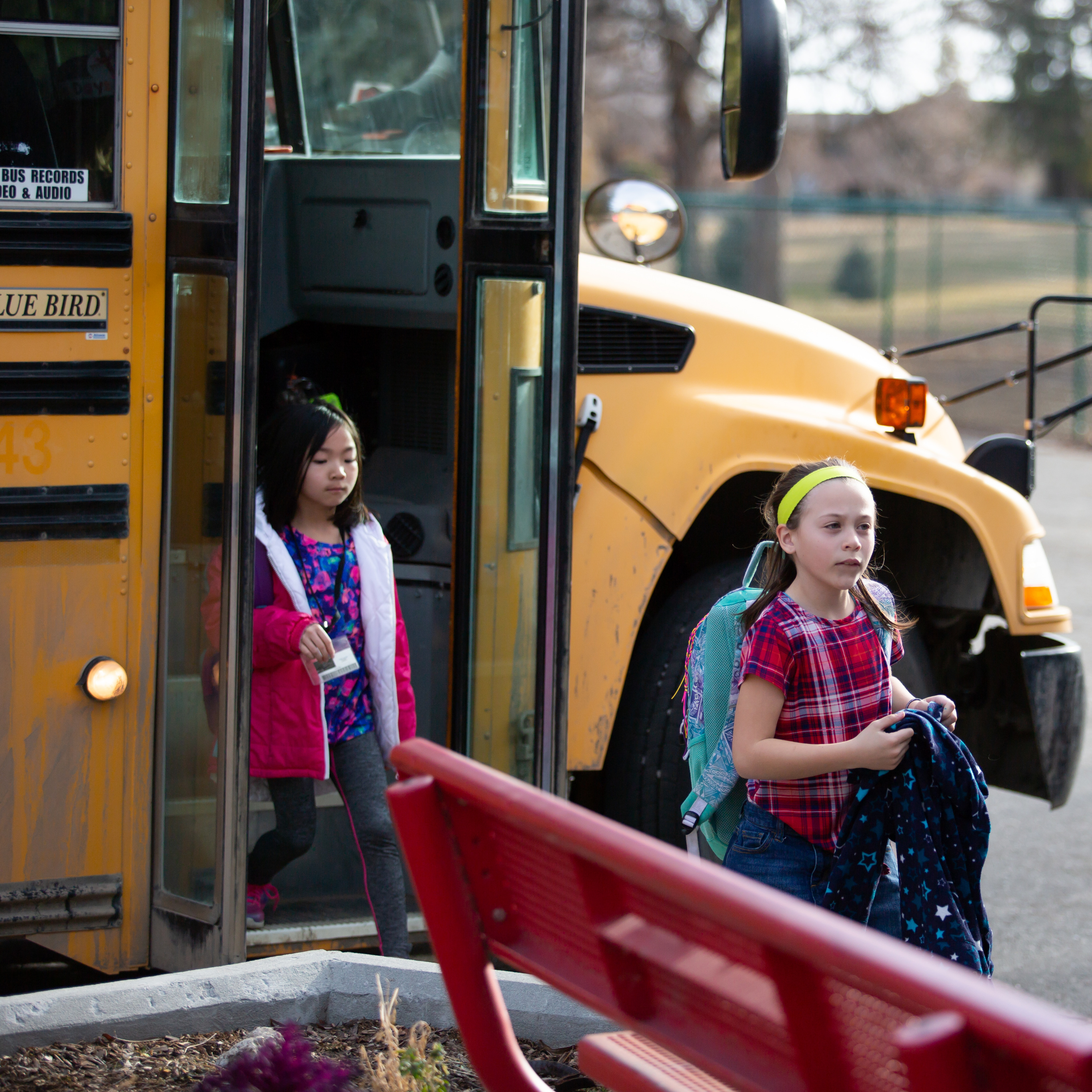 Two young children get off a yellow school bus holding backpacks. 
