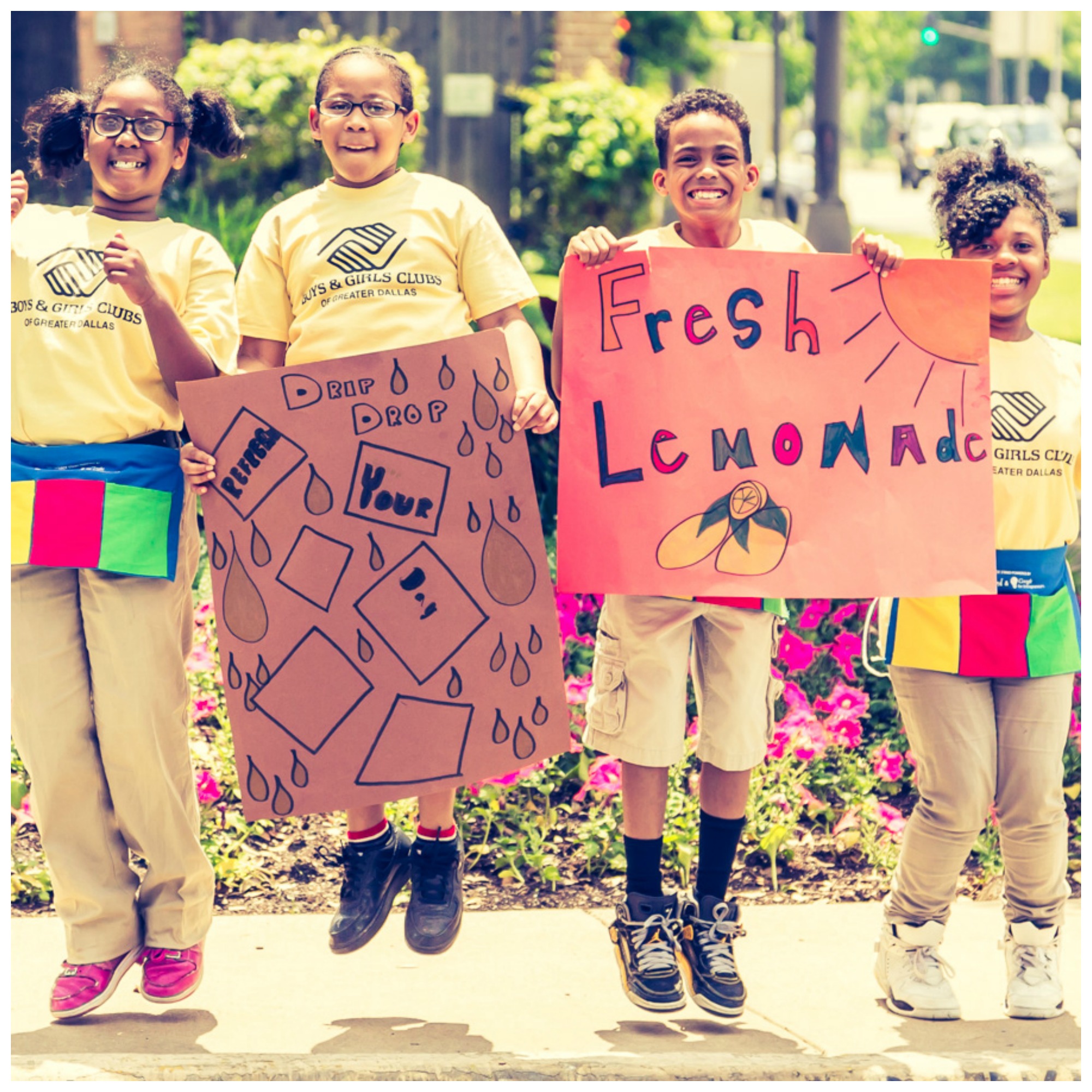 Four kids wearing yellow shirts that say "Boys and Girls Clubs" jump up and down in excitement while holding handmade signs that say "Drip Drop" and "Fresh Lemonade" 