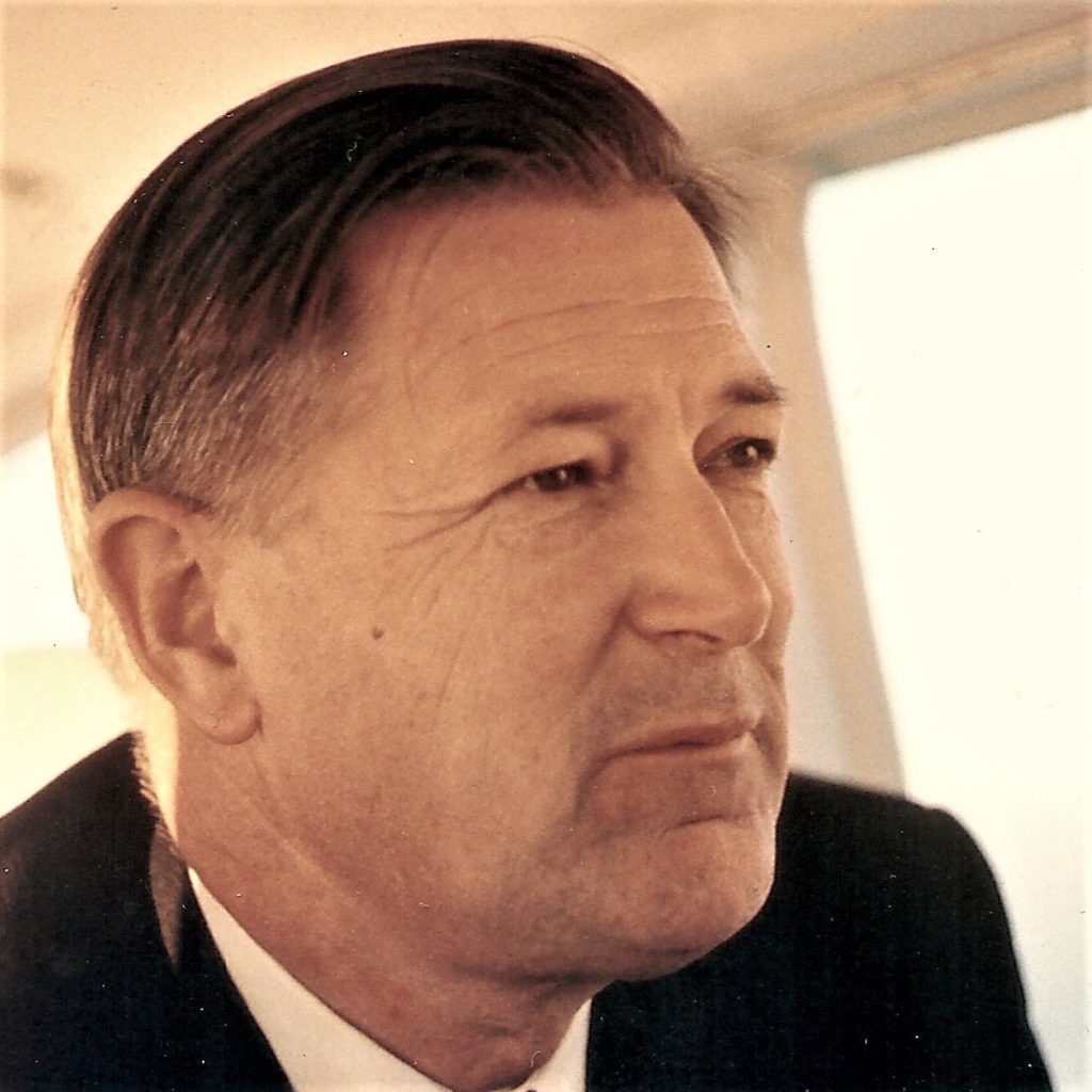 A close-up photo of Jack Murdock, a man with short dark hair wearing a black suit jacket, looking at something off camera. 