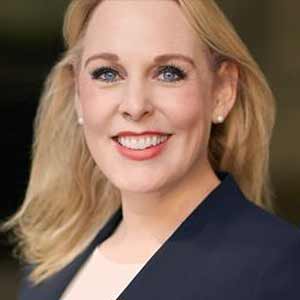 A woman with straight, blond, shoulder-length hair wearing a blue suit jacket smiles for the camera. 