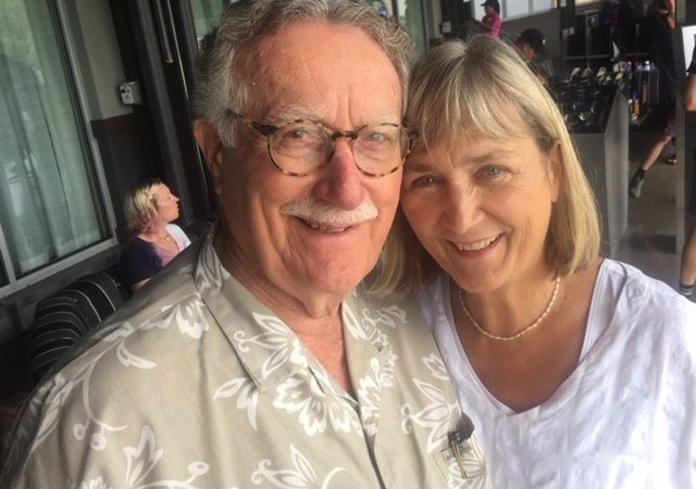 A man with gray hair and a mustache wearing a tan and white Hawaiian shirt and glasses smiles at the camera next to a woman with short blond hair and bangs wearing a white shirt and necklace. 