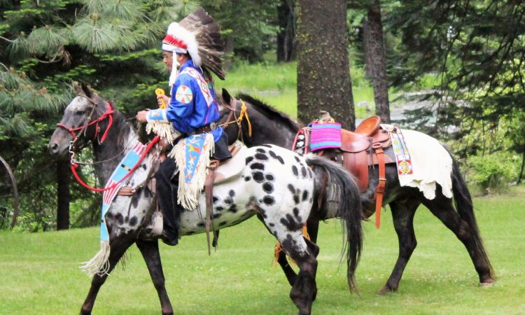 A person wearing a large headdress and traditional native attire rides a horse outside. A second horse with a saddle and bright blanket on its back is next to them. 