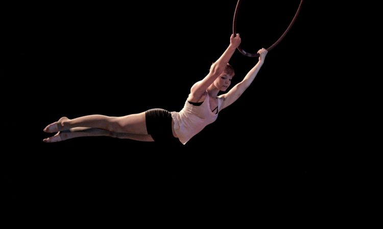 An acrobat swings from a hoop in front of a black background. 