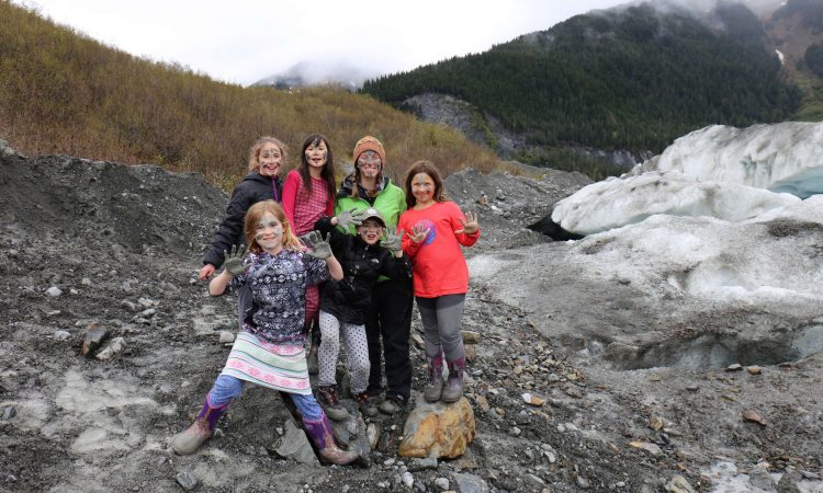 Five children smile at the camera and show off their muddy hands while standing outside surrounded by snow, mountains, and mud. 