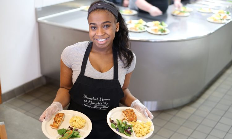 A woman wearing a gray shirt and black apron that says "Blanchet House of Hospitality" smiles at the camera while holding two plates of food inside a commercial kitchen.