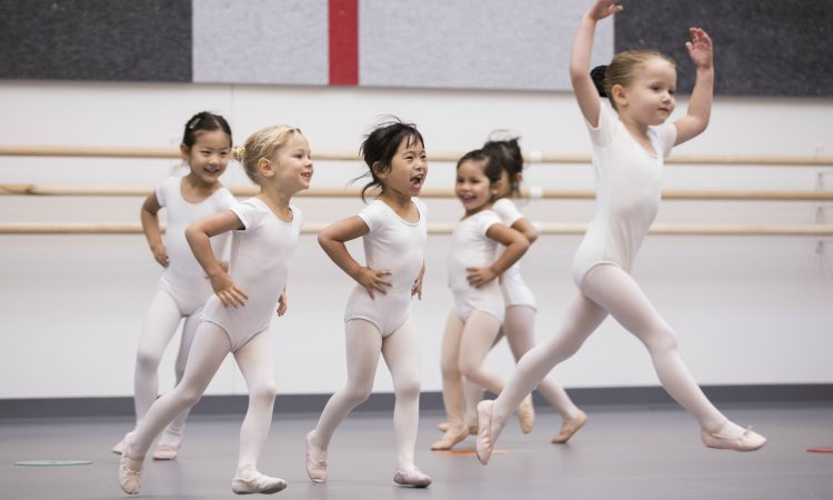 Six young girls in leotards and ballet slippers dance in a ballet studio.