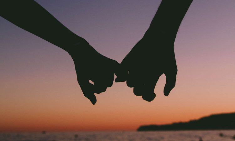 A silhouette of two hands linking pinky fingers with a sunset in the background. 