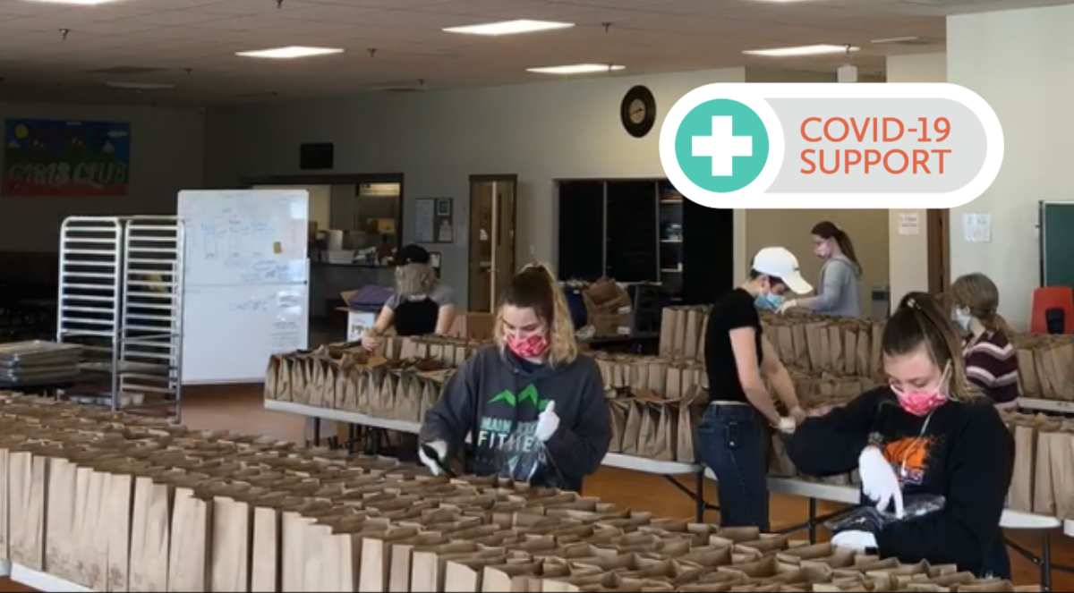 Volunteers fill brown paper bags with food and supplies inside a large room. Text overlay that says "COVID-19 SUPPORT"