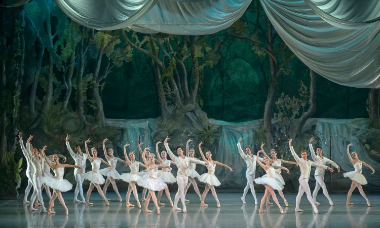 Ballerinas perform on a stage with trees in the stage backdrop. 