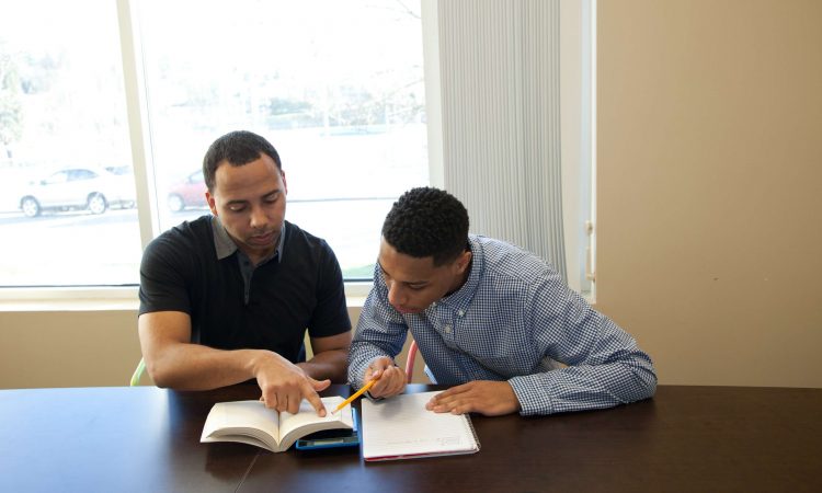 Two young men look at a book together while sitting at a table. 