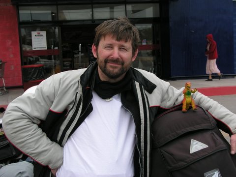 A man with brown hair and a beard smiles for the camera with his arm around a backpack. 