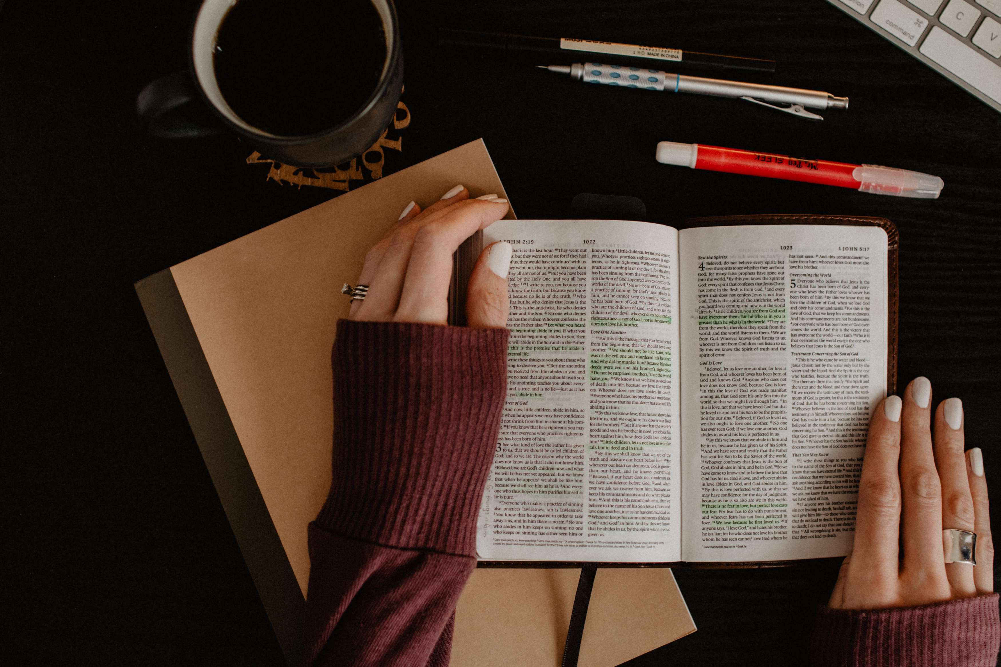 Two hands holding open a Bible on a table, with a notebook, pens, and coffee mug nearby. 